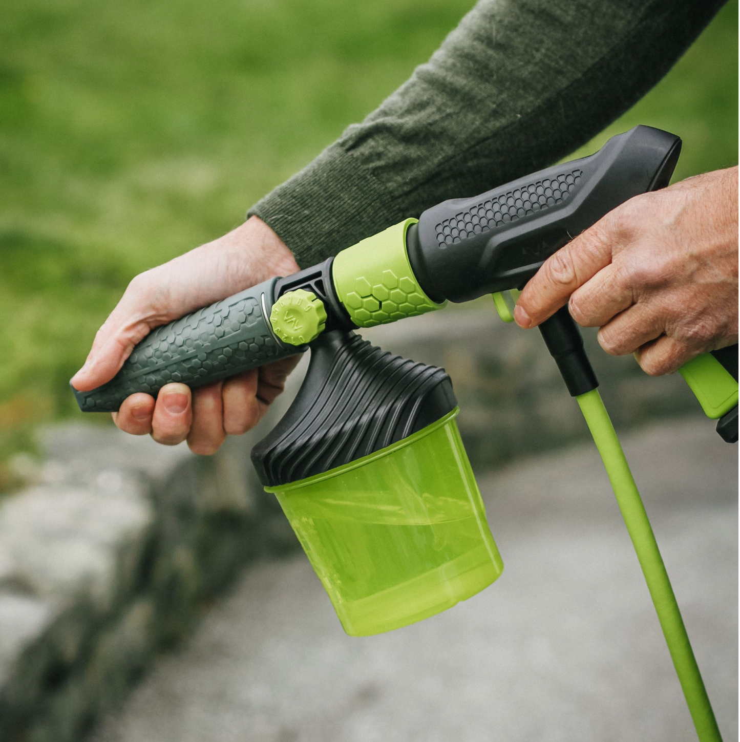 A person holding a translucent green soap container with a black lid, connected to a AVA zero force gun, with textured grips, shown outdoors on a paved surface with grass in the background.