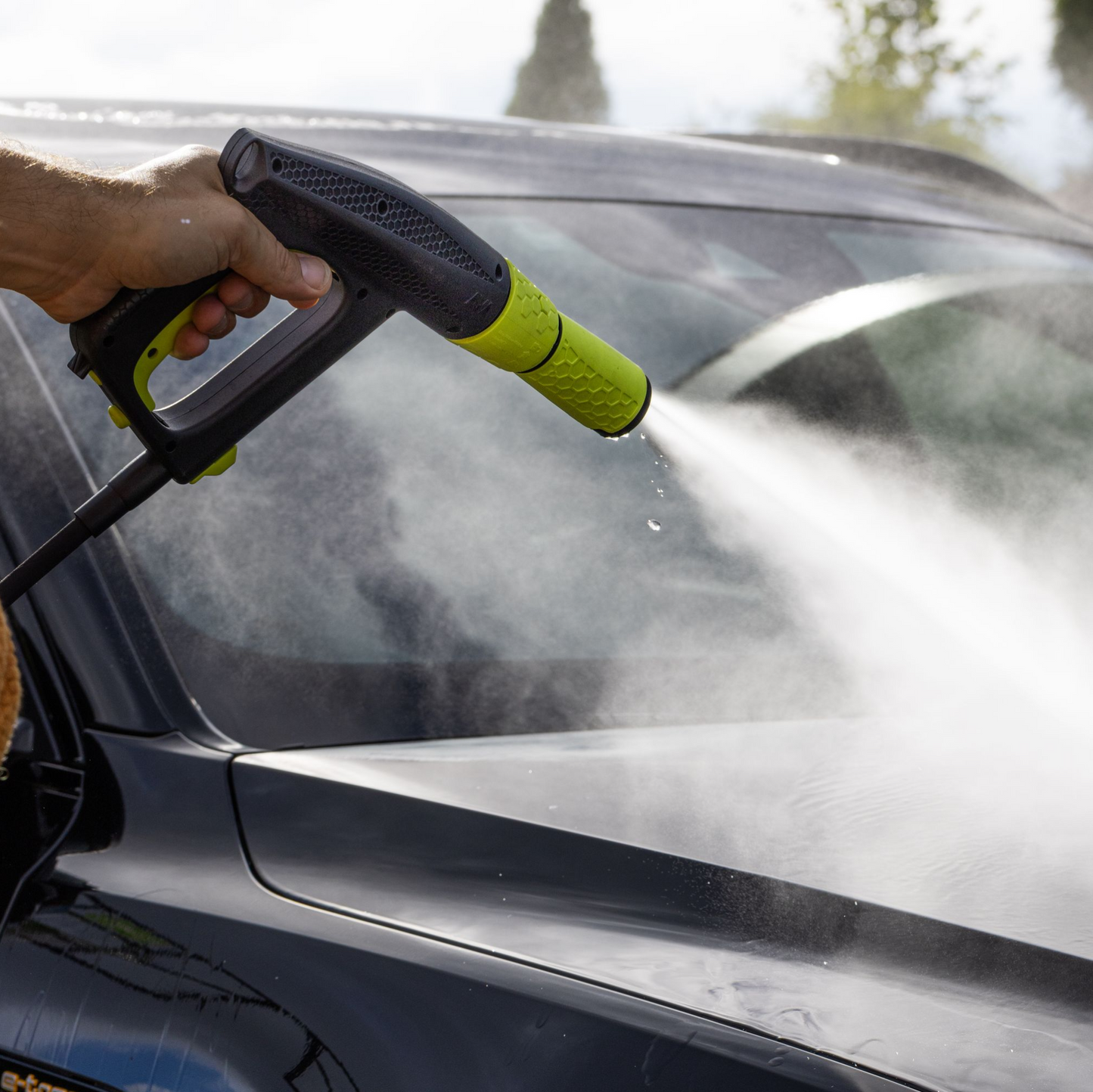 A person using a black and green high-pressure spray gun with an extra nozzle to wash a dark-colored car, with a strong jet of water directed at the car's windshield and hood.